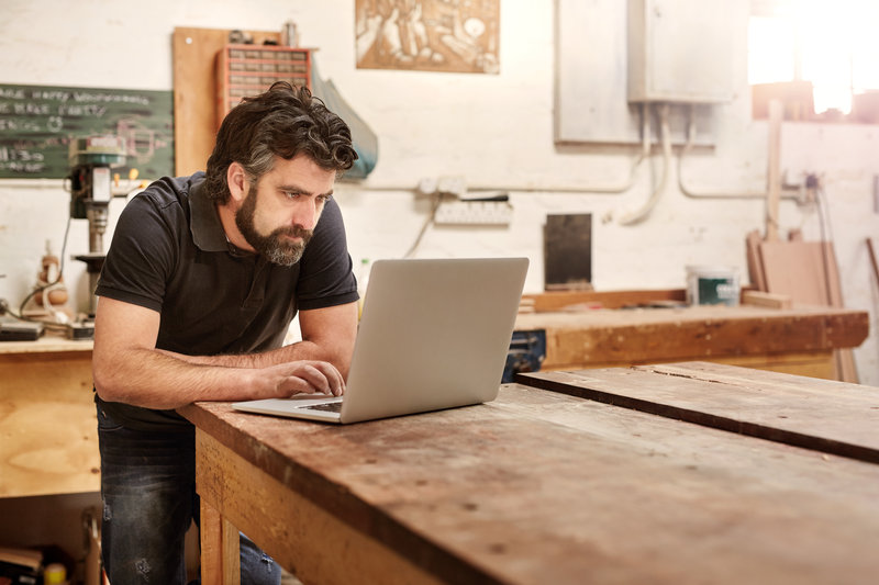 Bearded man who owns a small business, bending over at his work bench to type on his laptop, while working in his workshop and design studio