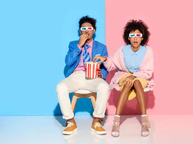 couple afroaméricain sur fond bleu et rose pastel qui regardent la télé en mangeant du pop corn